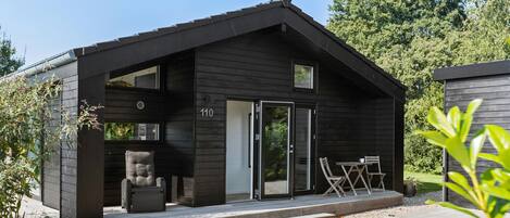 Una cabaña moderna con fachada oscura, puerta de vidrio corredera, un pequeño patio con mesa y sillas, y el número '110' en la pared.