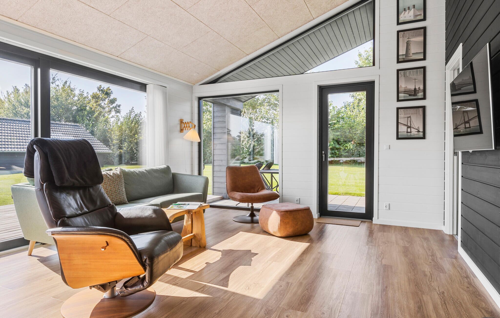 Un salon moderne avec un fauteuil en cuir, un canapé et une table basse. Des tableaux encadrés ornent le mur, et une grande fenêtre offre une vue sur l’extérieur.
