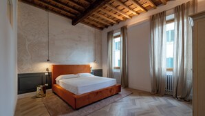 A bedroom with a wooden ceiling, a bed with white linens, a radiator, and a window with curtains. - House Bacco (Firenze)