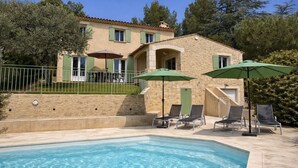 Ein Poolbereich mit Liegestühlen und Sonnenschirmen, ein Gebäude mit Steinbau und einem Balkon sowie ein klares, blaues Schwimmbecken.
