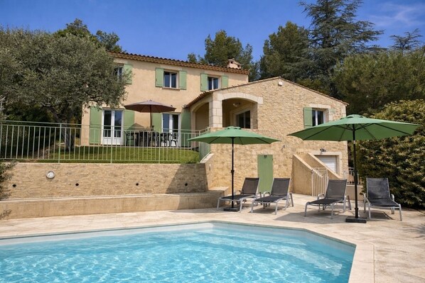A poolside area with lounge chairs and umbrellas, a stone building with a balcony, and a clear blue pool.