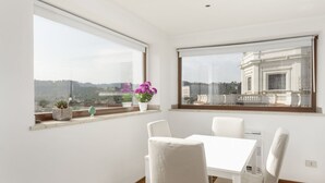 A modern room with a white table and chairs, large windows with views of buildings and greenery, and potted plants on the windowsill. - LaDolcevita - Ponte Milvio Stunning View Penthouse (Roma)