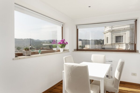 Une pièce moderne avec une table et des chaises blanches, de grandes fenêtres offrant une vue sur les bâtiments et la verdure, et des plantes en pot sur le rebord de la fenêtre.