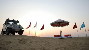A desert landscape with a dune buggy parked on the sand, flags, and a beach umbrella. - Rajasthan Desert Camp  (Jaisalmer)