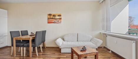 A small, well-lit room with a wooden dining table and chairs, a sofa, and a small wooden table with decorative items.
