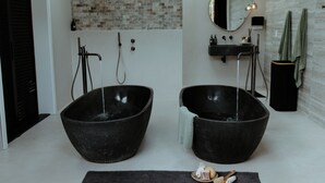 Une salle de bain moderne avec deux grands lavabos noirs, un mur au motif de briques, un miroir rond et un petit coin lavabo.