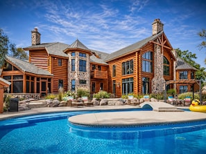 A large log cabin with a swimming pool, stone chimneys, and a well-maintained garden.
