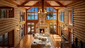 A log cabin interior with a stone fireplace, wooden floors, and large windows offering views of a lake.