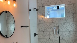 Un baño moderno con un diseño de baldosas geométricas, un espejo grande, un lavamanos con grifo negro, un inodoro y una ducha con cabezal de ducha de mano.