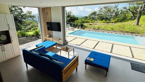 Ein modernes Wohnzimmer mit einer großen Couch und einem Couchtisch, einem an der Wand befestigten Fernseher und Blick auf einen Swimmingpool sowie üppige Grünflächen.