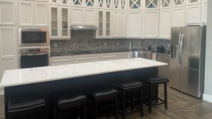 Une cuisine moderne avec des meubles blancs, un grand îlot central doté de tabourets, des appareils en acier inoxydable et un pare-feu décoratif.