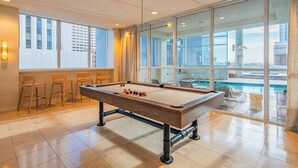 A pool table in a room with large windows, a view of a cityscape, and a pool area visible through the glass. - ROVE LIVING/KL - 44 Monroe (Phoenix)