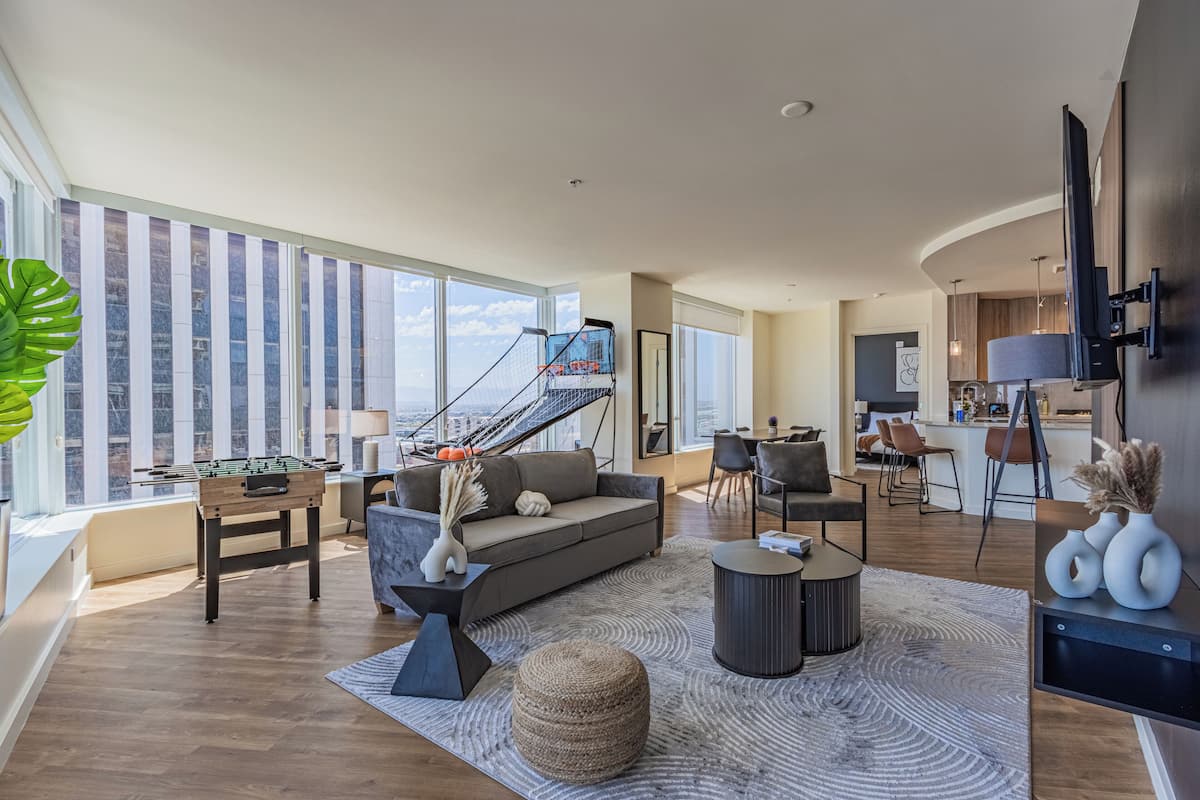 A spacious living area with a sofa, coffee table, and a foosball table. There is a staircase leading to an upper level, and a kitchen area is visible in the background.