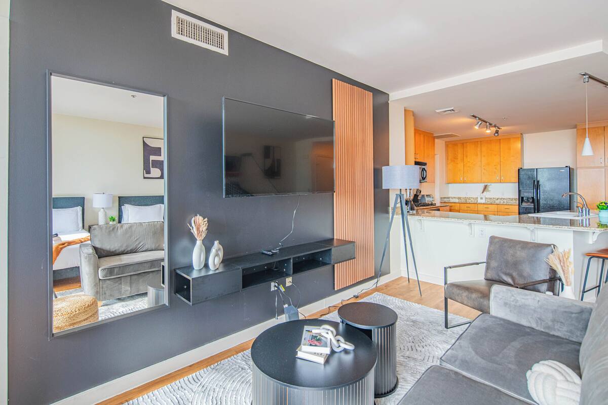 A modern living room with a flat-screen TV, sofa, coffee table, and a kitchen area with wooden cabinets and a refrigerator.