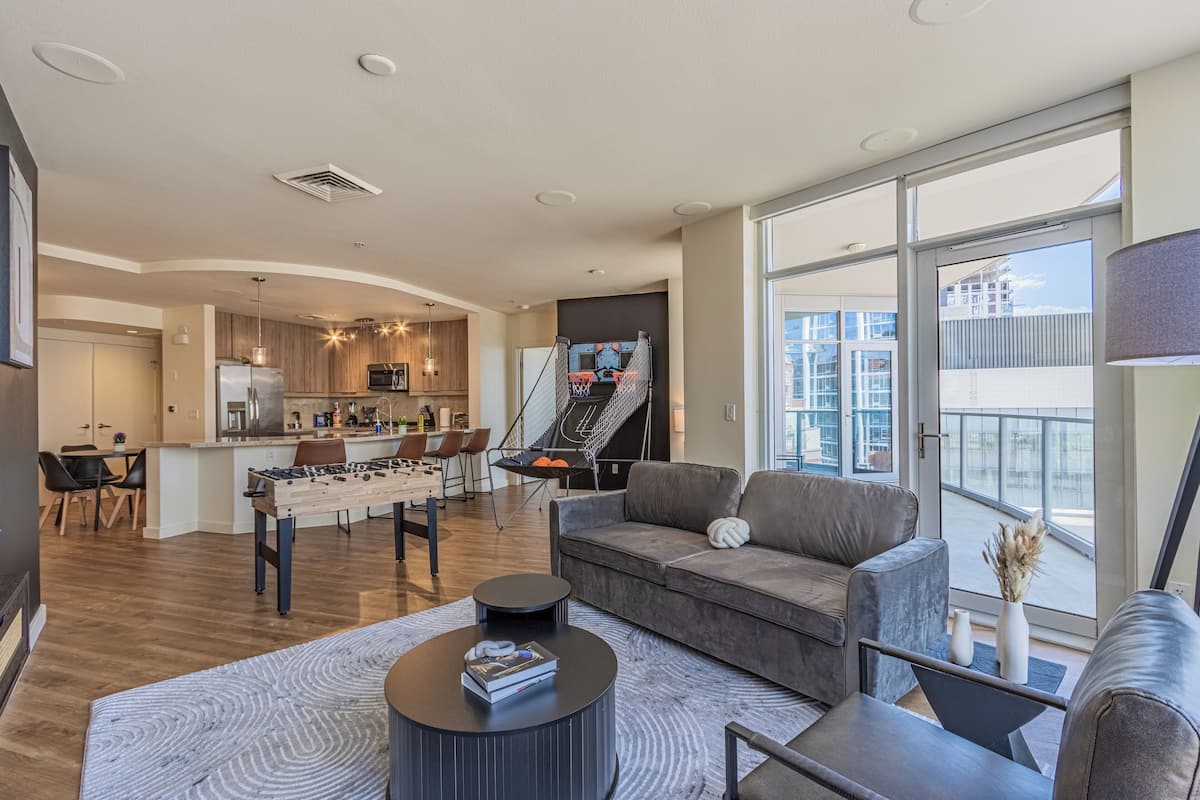 A modern living room with a grey sofa, a coffee table, and a foosball table. There is a kitchen area with a dining table and chairs, and a large window offering a view of the city.