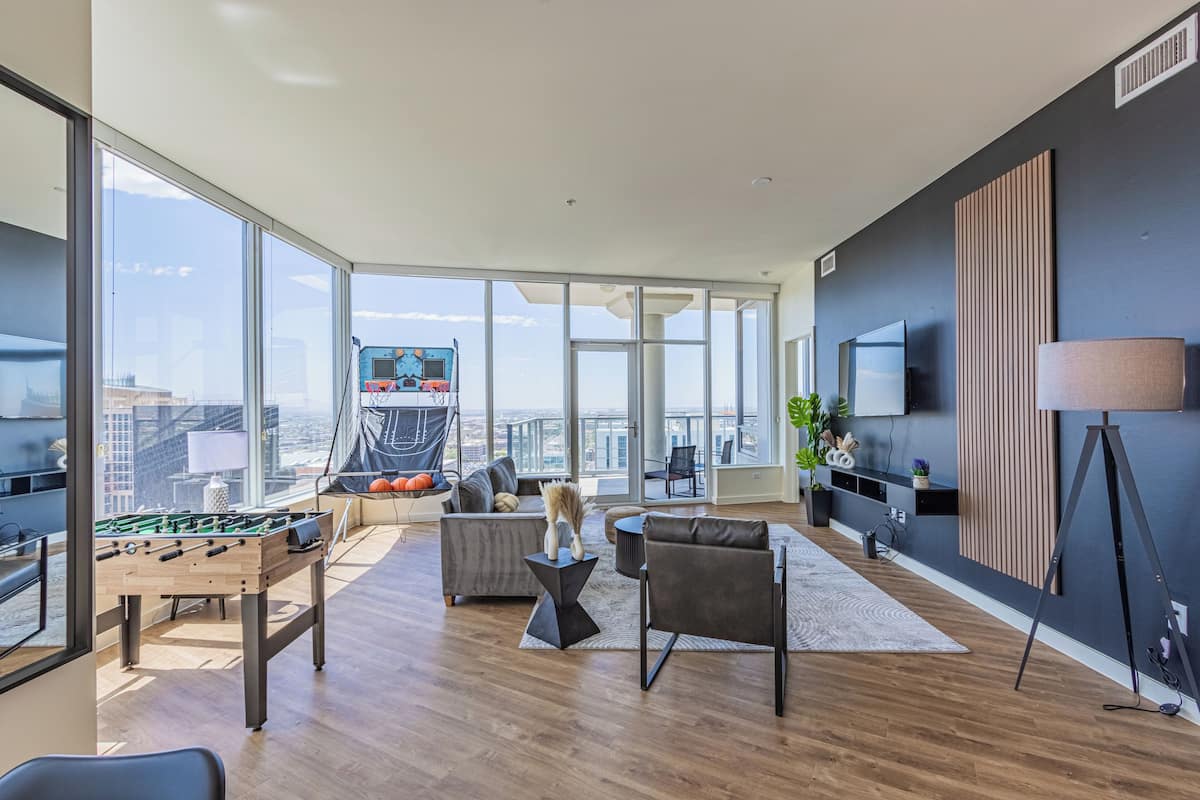 A spacious living area with a foosball table, a grey sofa, and a large window offering a city view.