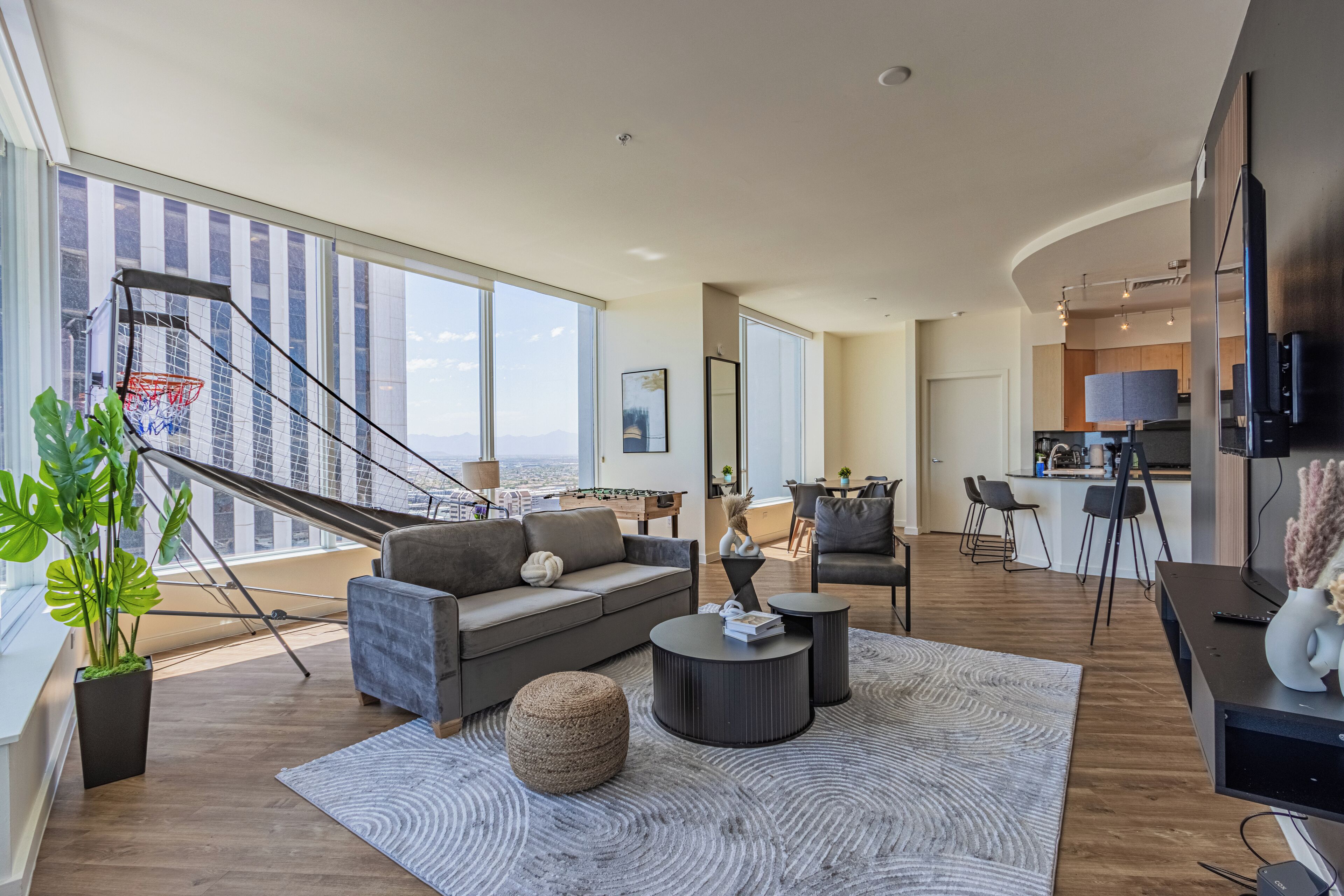 A modern living room with a sofa, coffee table, and ottoman, a staircase leading to a balcony, and a kitchen area visible in the background.
