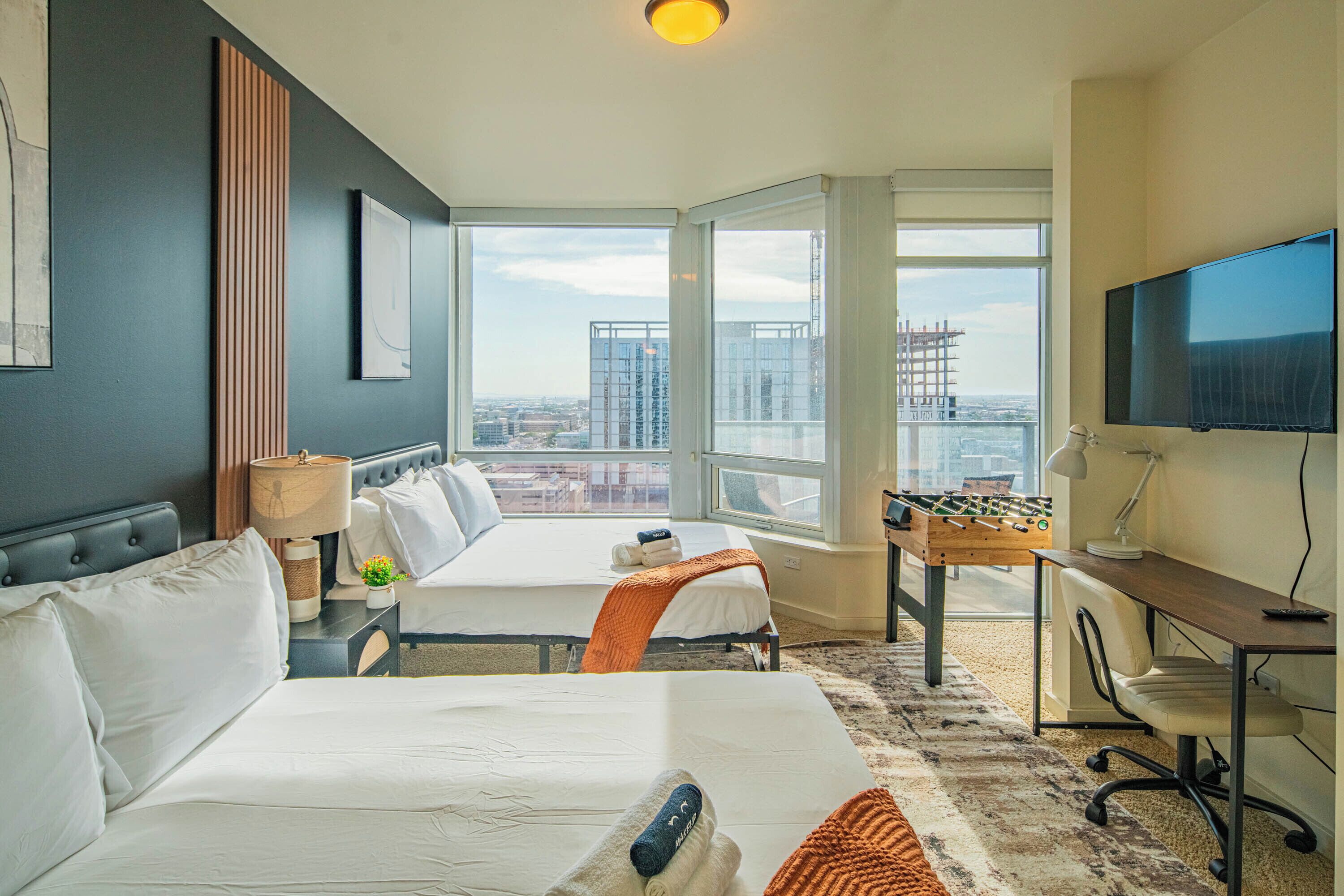 A hotel room with two beds, a foosball table, a desk with a computer, a TV, and a large window with a city view.