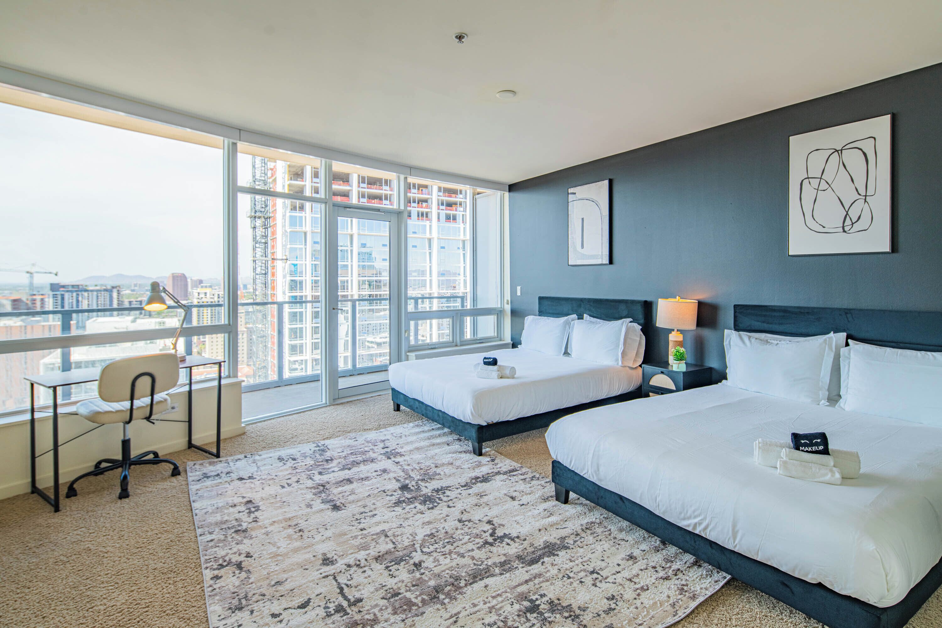 A modern hotel room with two beds, a desk, and a large window offering a city view.