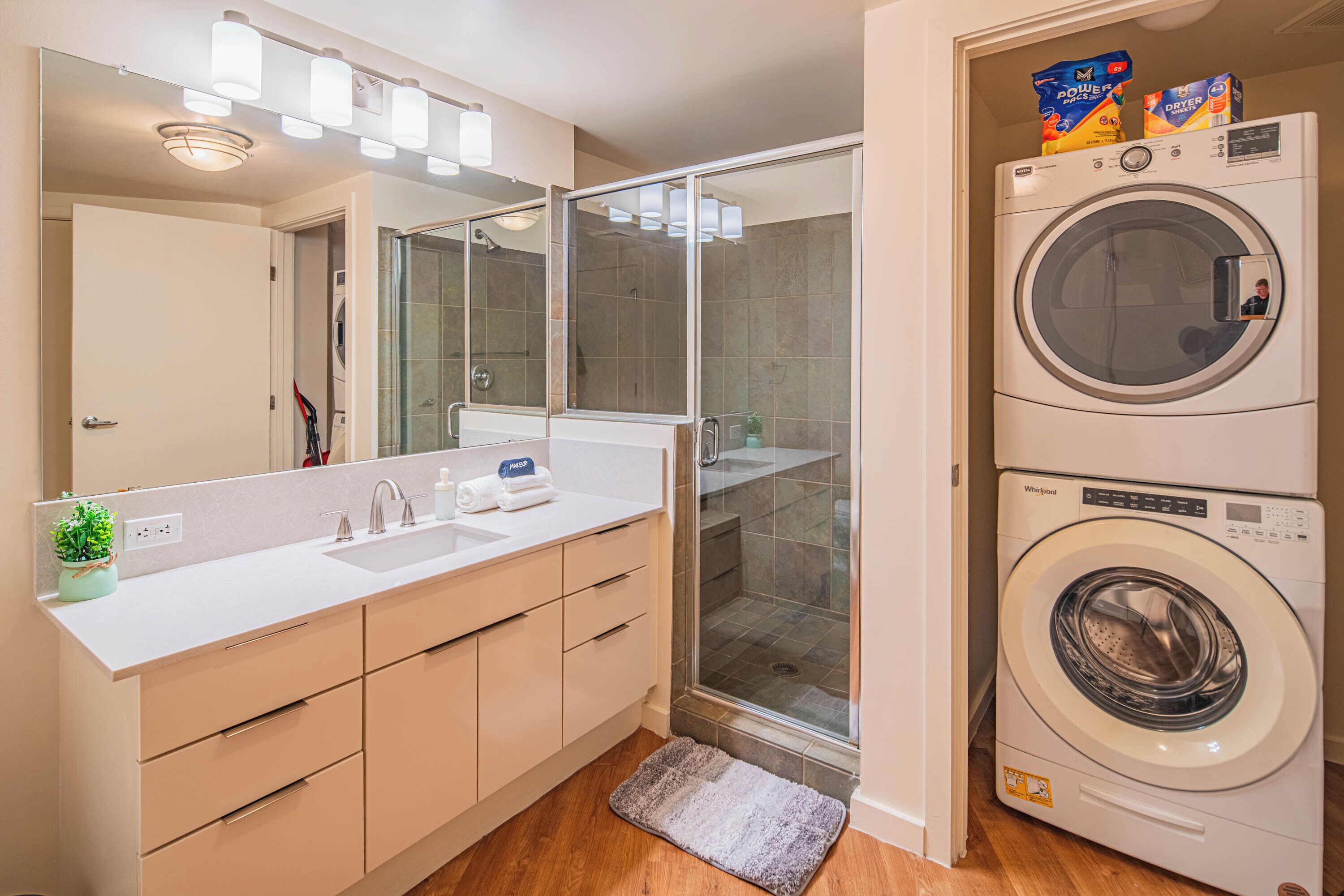 A modern bathroom with a large mirror, a shower, a built-in washing machine, and a white vanity.