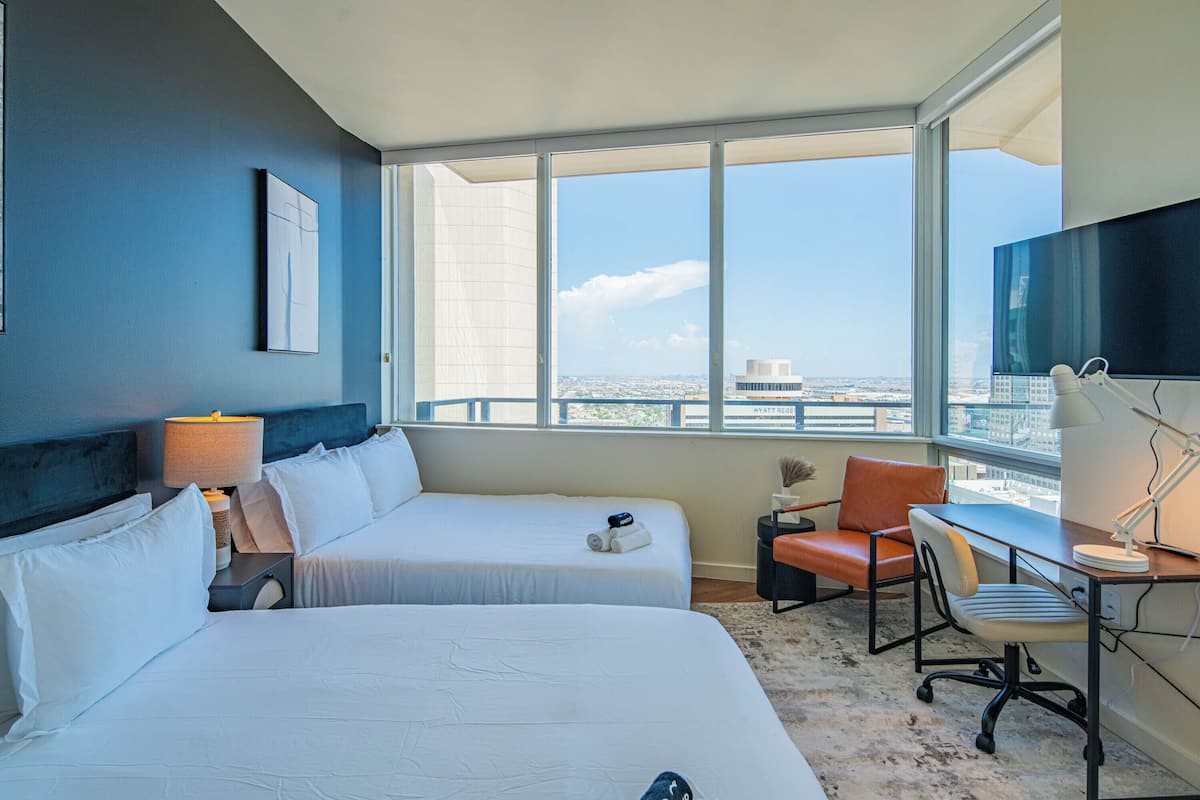 A hotel room with two beds, a desk, a chair, and a large window offering a city view.