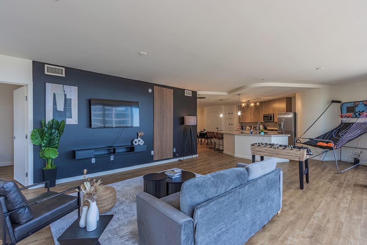 A modern living room with a grey sofa, a coffee table, and a TV mounted on the wall. There is a kitchen area in the background with a dining table and chairs.