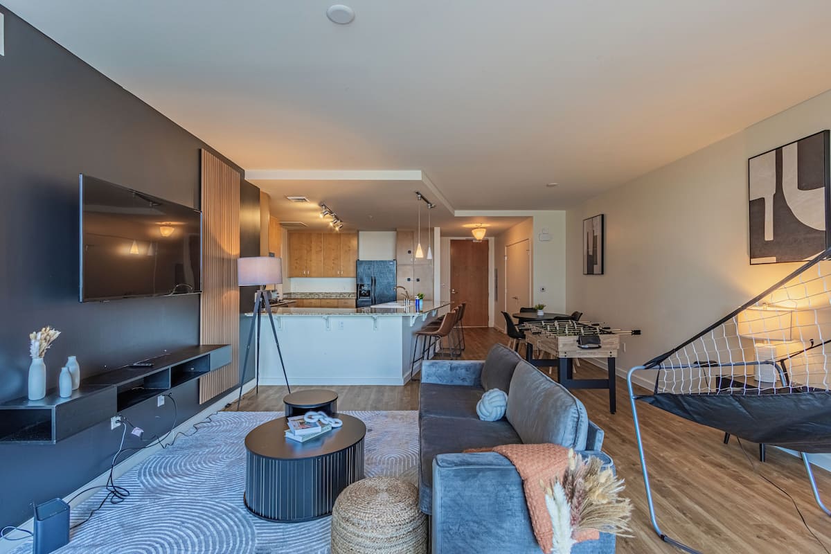A modern living room with a sofa, coffee table, and TV. There is a kitchen in the background with a dining area.