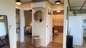 A compact living space with a wooden floor, a white refrigerator, a small trash bin, and a bathroom with a shower curtain. - Mountain and Water View at Charming 2-bedroom Retreat in Sequim (Sequim)