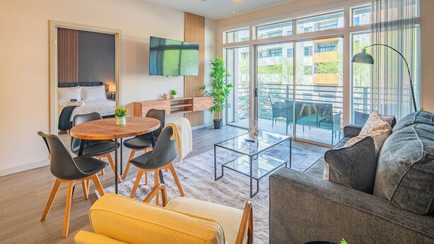 Un salon moderne comprenant un canapé, une table à manger avec des chaises, un téléviseur fixé au mur et de grandes fenêtres offrant une vue sur l’extérieur.