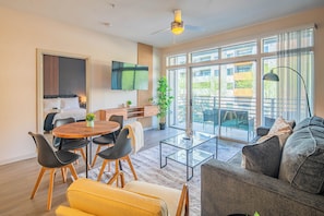 Una sala de estar moderna con un sofá, una mesa de comedor con sillas, un televisor montado en la pared y grandes ventanas con vistas al exterior.