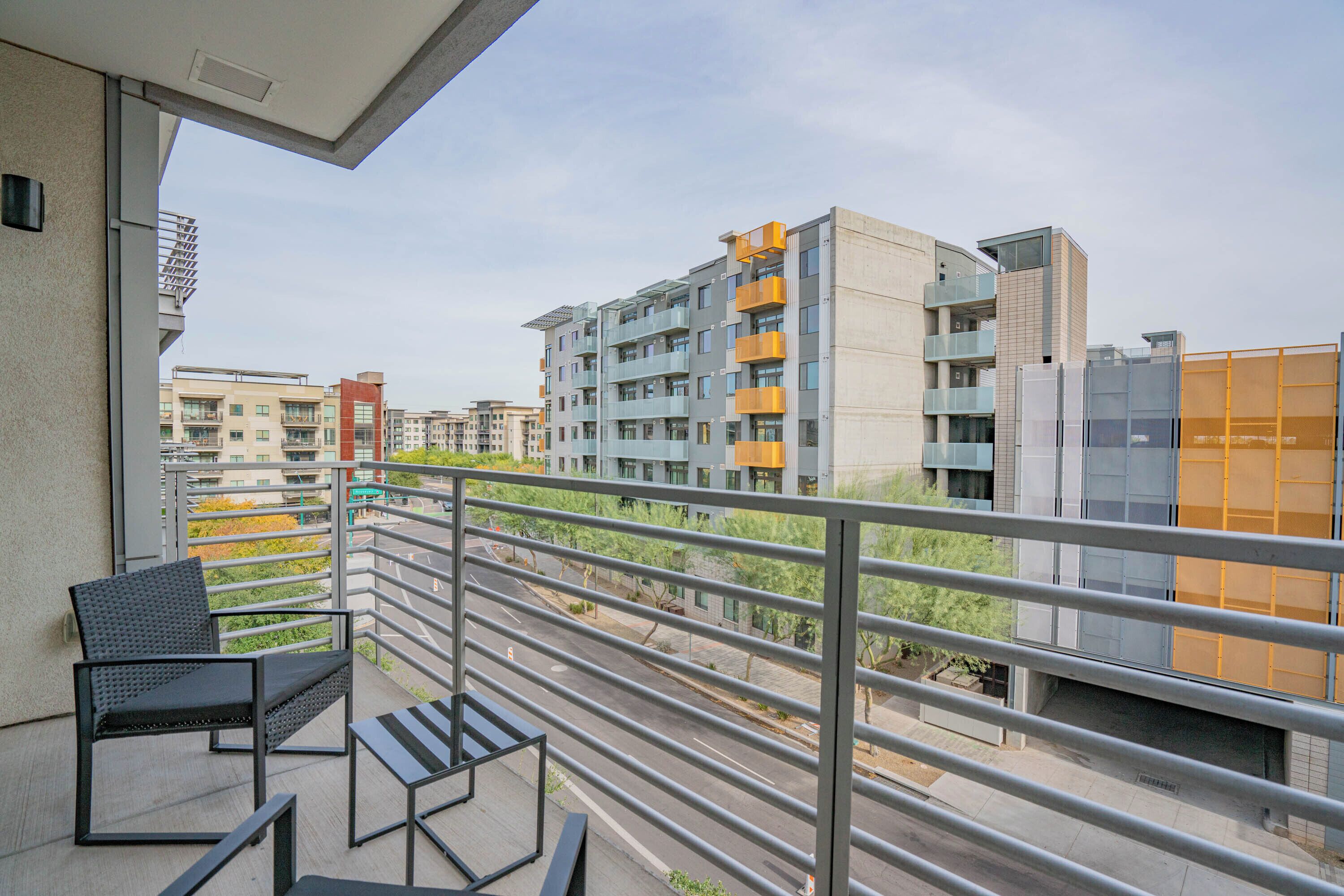A balcony with a chair and table, overlooking a street with buildings and greenery.