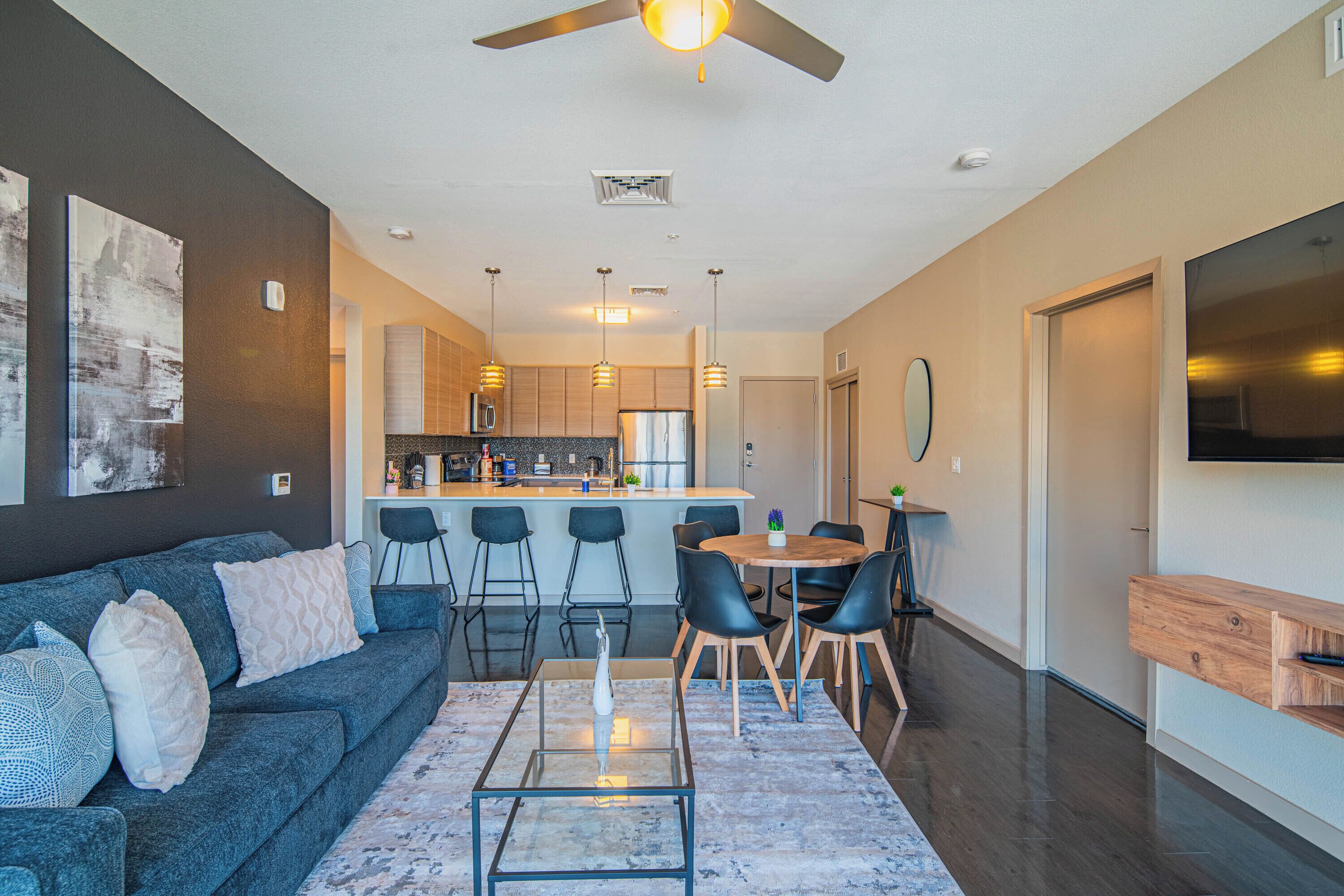 A modern living room with a sofa, coffee table, and chairs. A kitchen with bar stools is visible in the background.