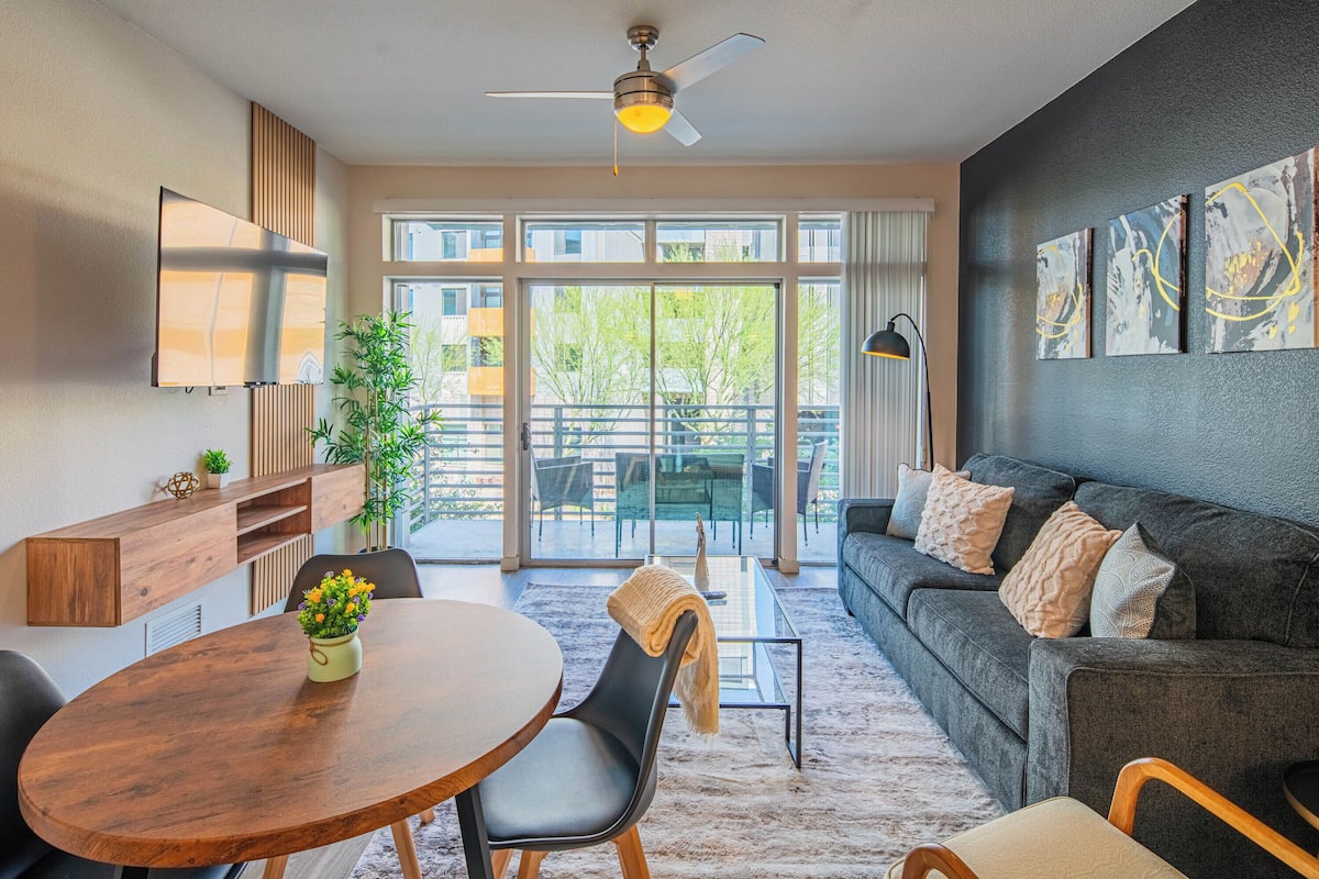 Ein modernes Wohnzimmer mit einem dunkelgrauen Sofa, einem Holztisch und einem Esstisch mit Stühlen. Es gibt einen Deckenventilator, ein großes Fenster mit Blick ins Freie und Kunstwerke an der Wand.