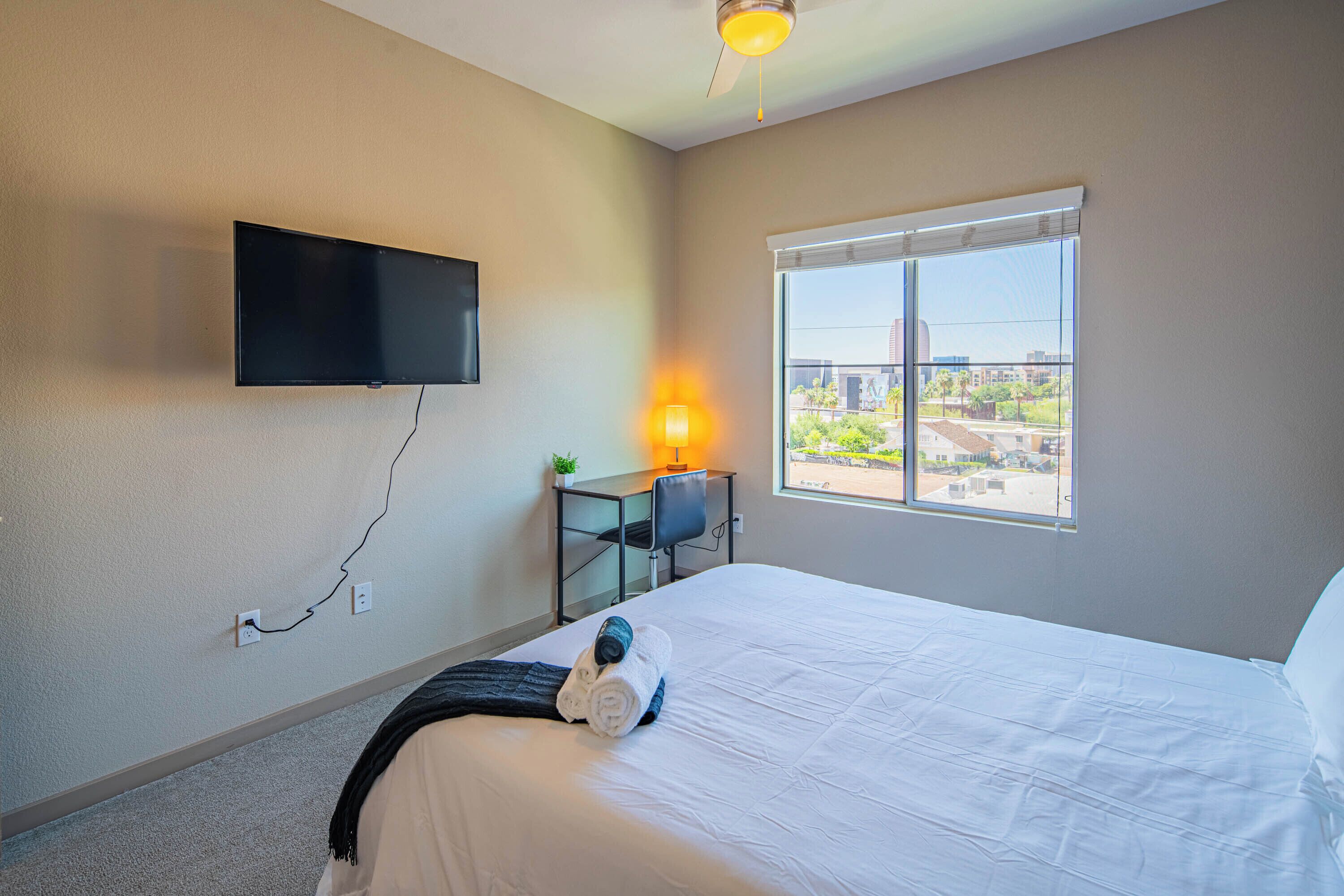 A hotel room with a bed, a desk with a chair, a TV mounted on the wall, and a window with a view of the city.