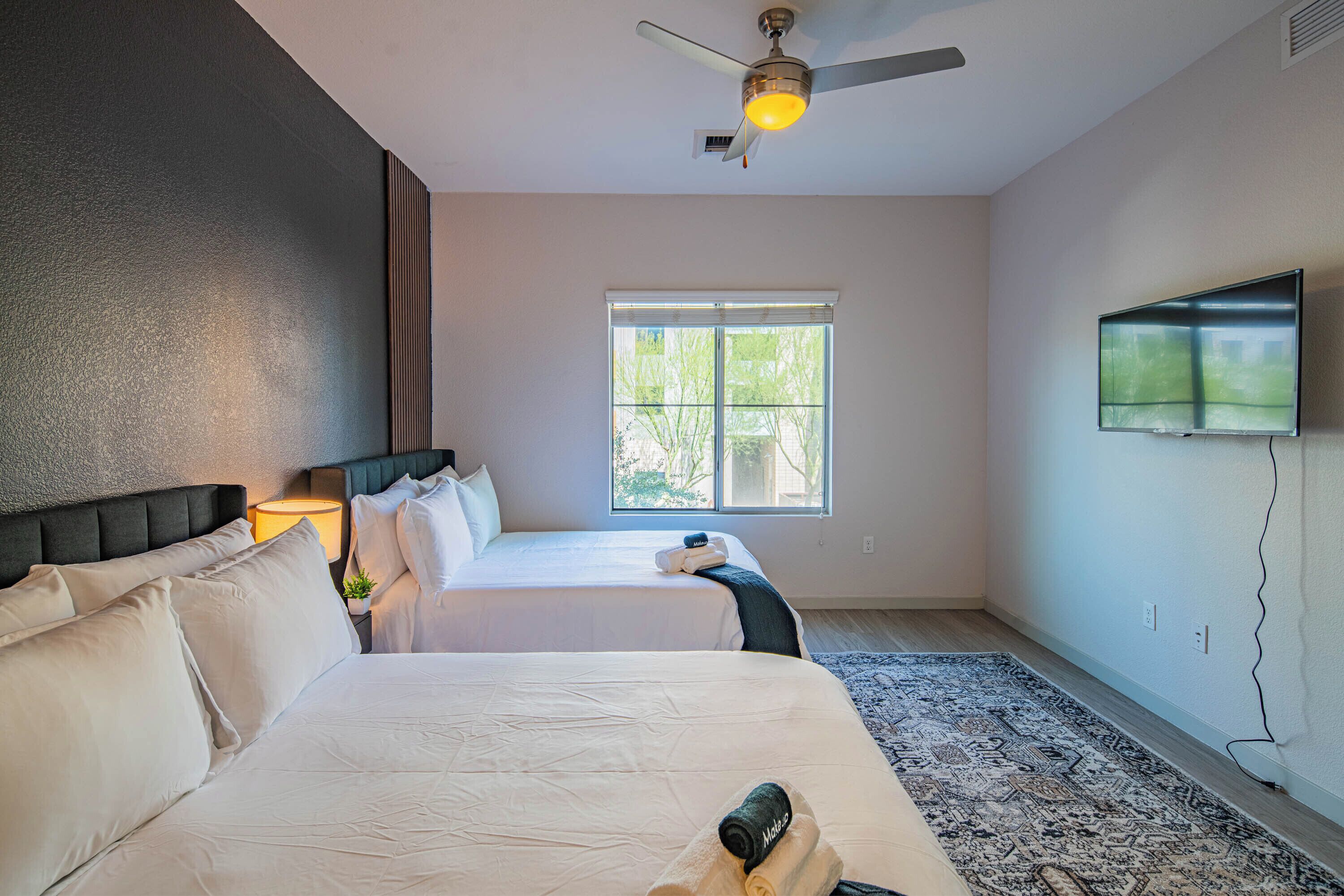 A hotel room with two beds, a ceiling fan, a TV mounted on the wall, and a window with a view of trees.
