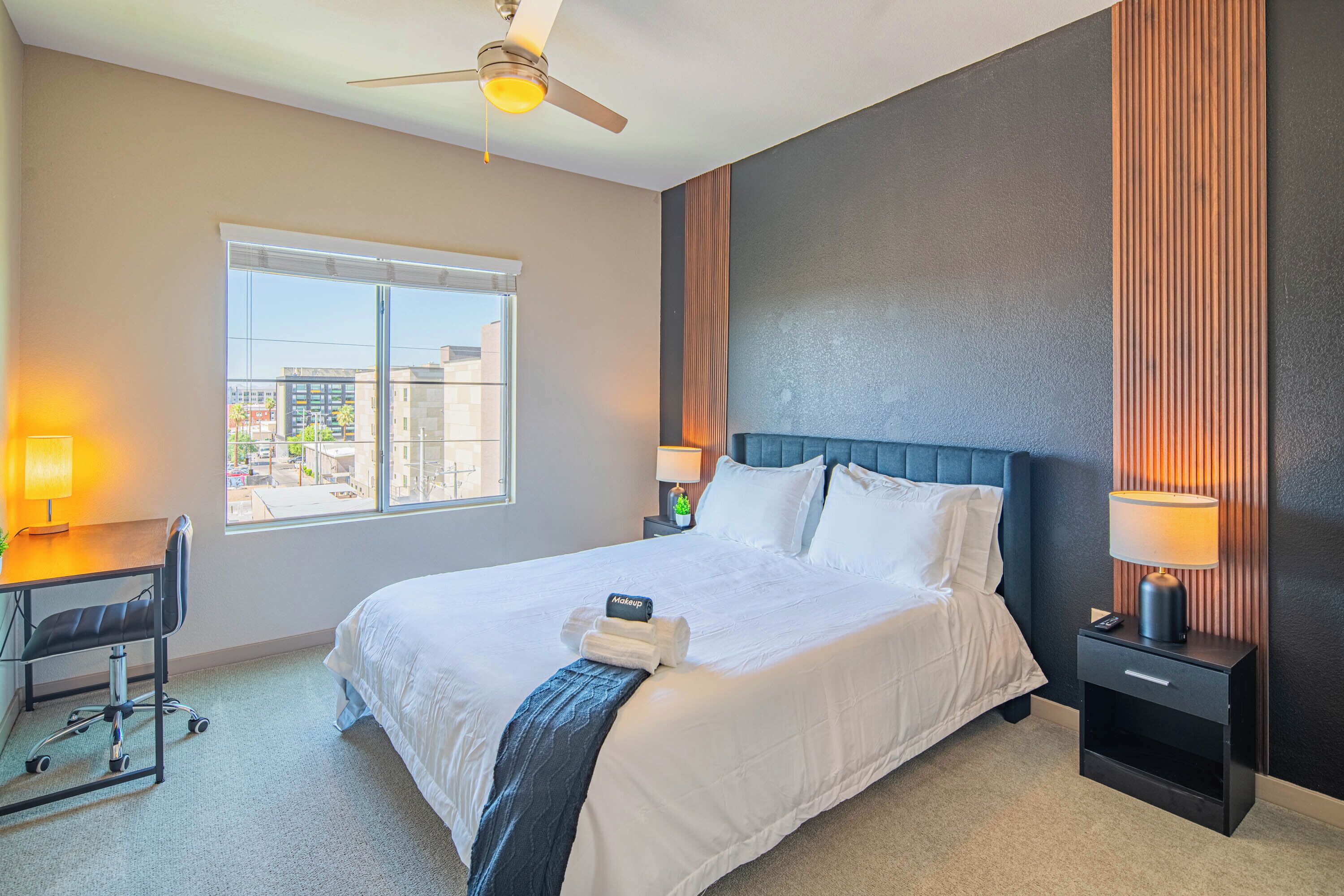 A hotel room with a large bed, a desk with a chair, a nightstand with a lamp, and a ceiling fan.