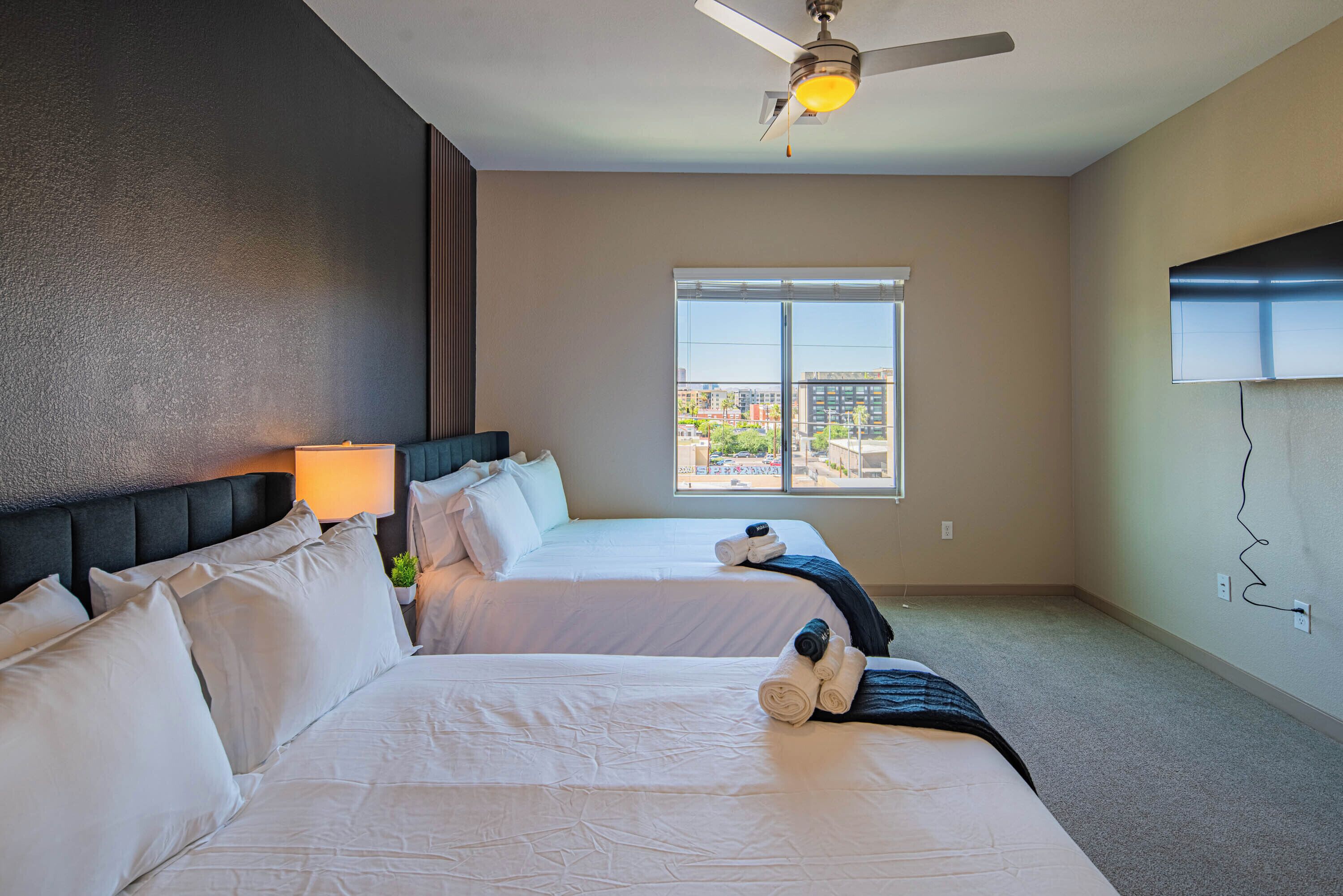 Habitación de hotel con dos camas, ventilador de techo, ventana con vistas a la ciudad y televisión montada en la pared.