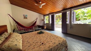 A bedroom with a hammock, a bed with floral bedding, a ceiling fan, and a view of greenery through open doors.