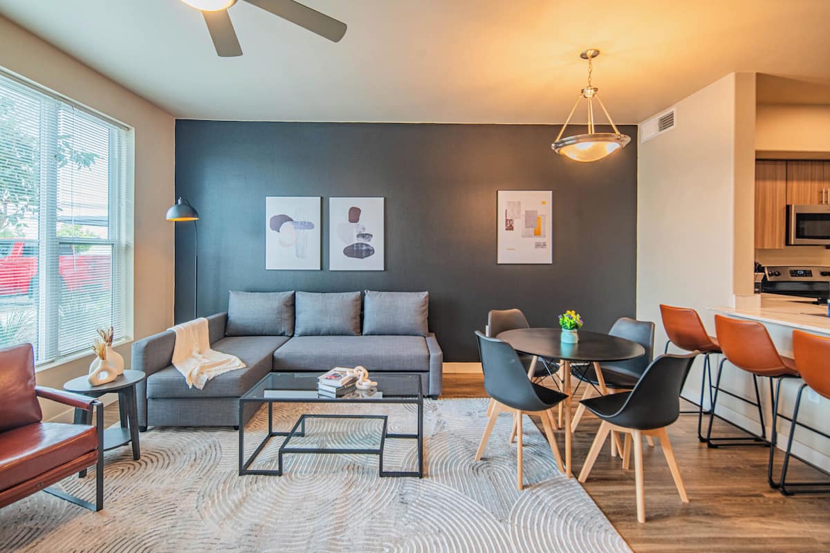 A modern living room with a grey sofa, coffee table, and dining area with chairs. There is a kitchen in the background with a microwave and cabinets.