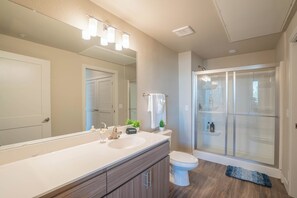 A modern bathroom with a large mirror, dual sinks, a toilet, and a glass-enclosed shower. - ROVE LIVING/KL - V on Broadway (Tempe)