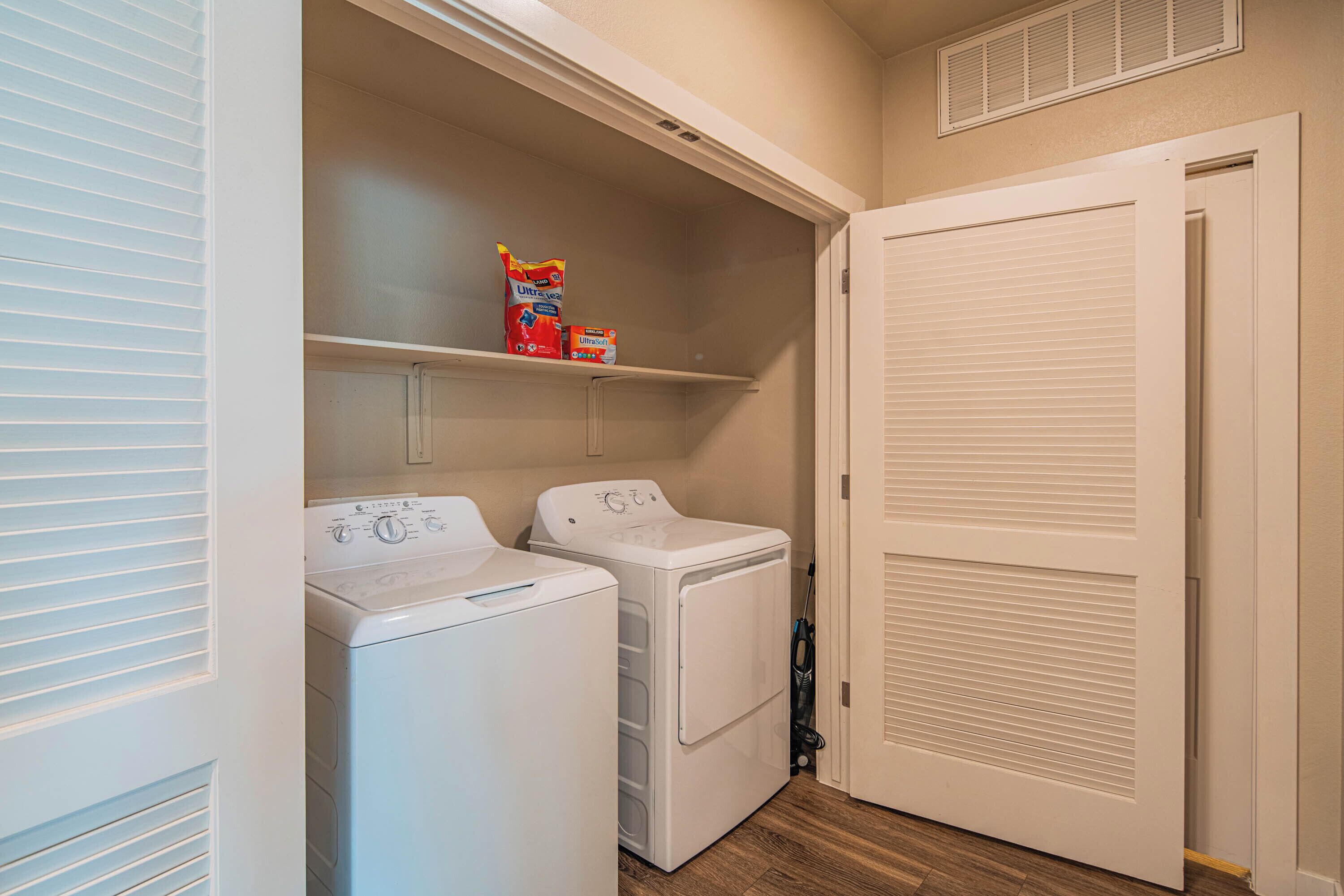 A laundry room with two front-loading washing machines, a shelf with laundry detergent, and a closed closet door.
