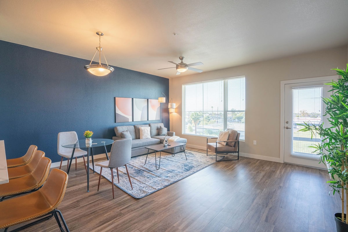 A modern living room with a blue accent wall, a ceiling fan, a dining area with chairs and a table, and a large window with blinds.