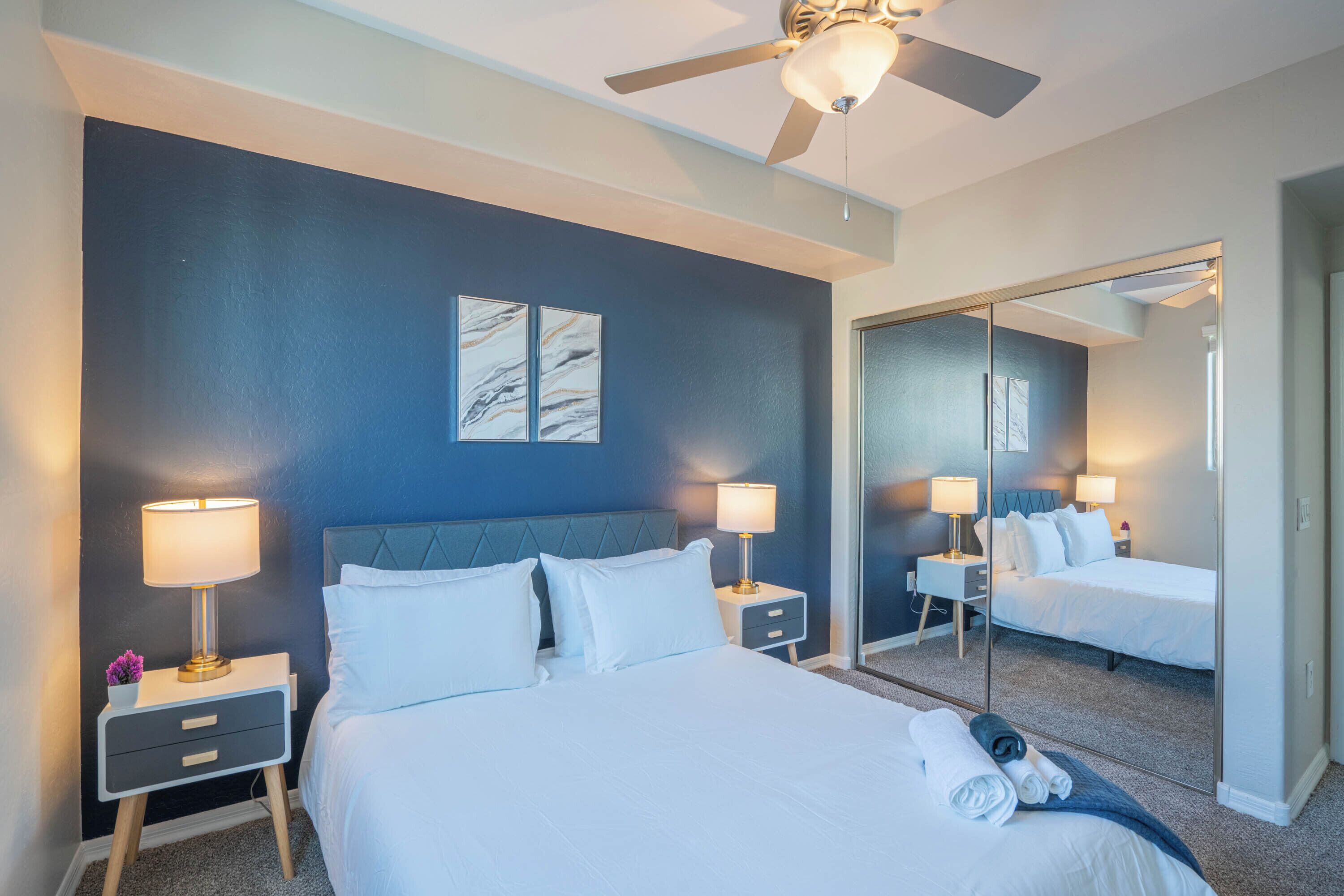 A hotel room with a large bed, two bedside tables with lamps, a ceiling fan, and a large mirror reflecting the room.