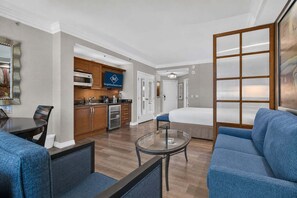 Habitación de hotel con kitchenette, una cama, una zona de estar con mesa de vidrio y un ventanal grande.