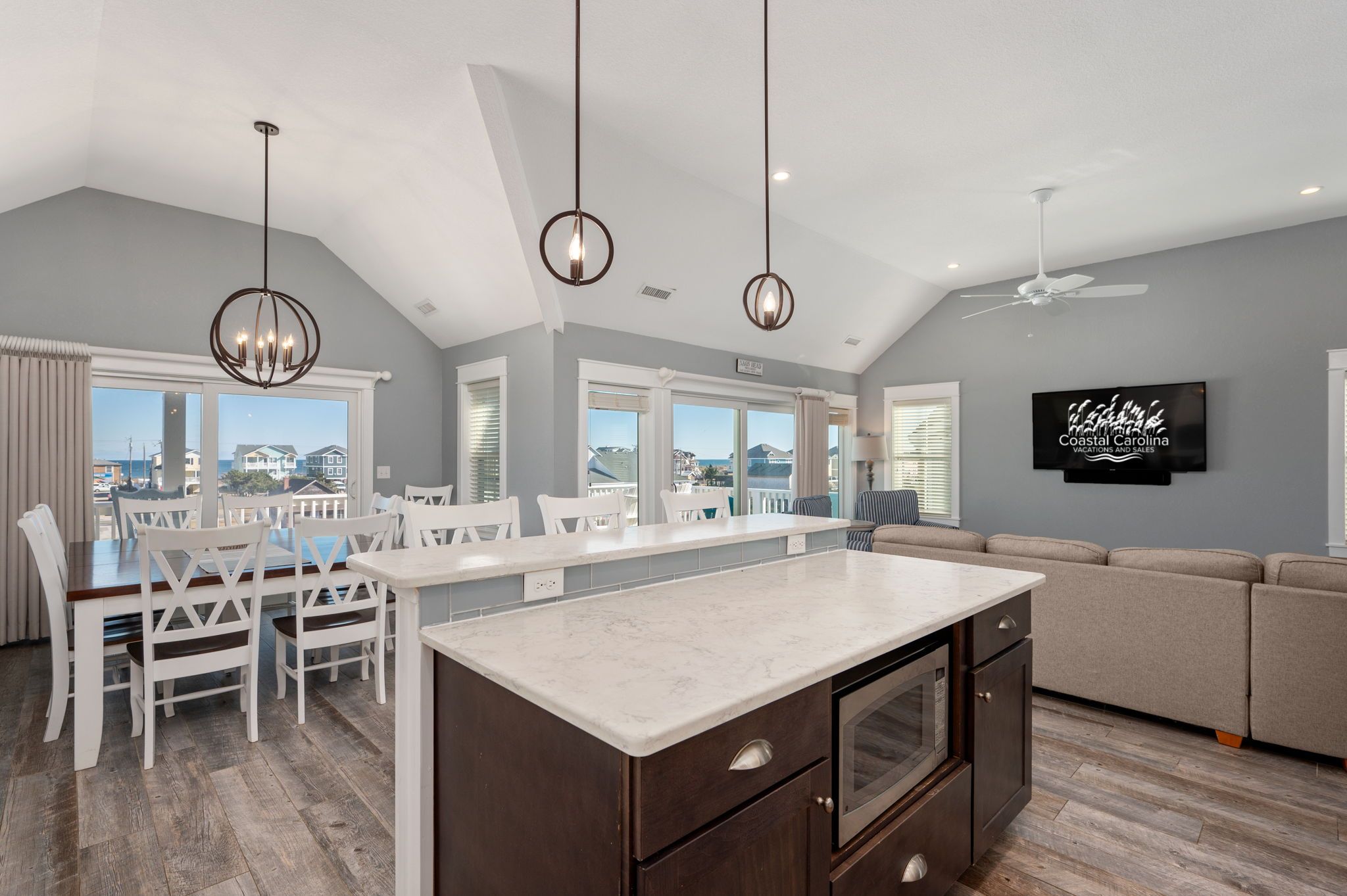 A spacious living area with a kitchen island, dining table, and seating, featuring large windows and a ceiling fan.