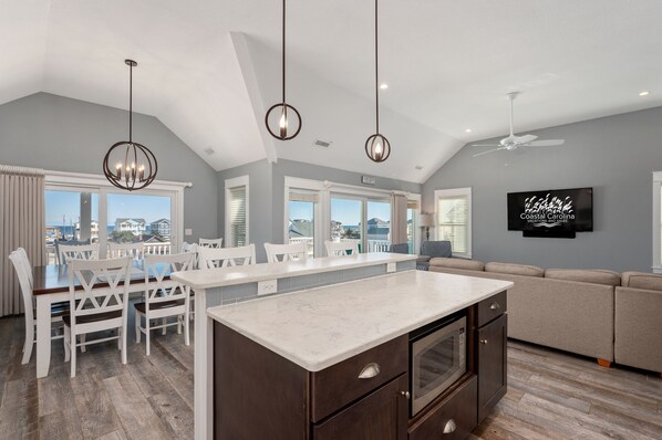 A spacious living area with a kitchen island, dining table, and seating, featuring large windows and a ceiling fan.