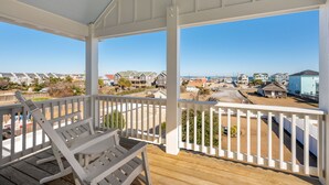 Uma varanda coberta com grades brancas, um deck de madeira e vista para uma área residencial com casas e uma praia ao fundo.