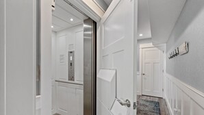 Um corredor com paredes brancas, piso de madeira e uma porta que dá para um ambiente com elevador de aço inoxidável.