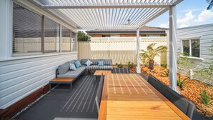 A covered patio with a wooden table, a gray sofa with cushions, and a white fence. - Summer Breeze Forster
The Perfect Family Getaway on the Mid North Coast of NSW (Forster)