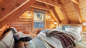 Un dormitorio acogedor en el ático, con techo de madera, un ventanal grande, una cama con sábanas blancas, un banco con mantas y una mesita de noche con lámpara.